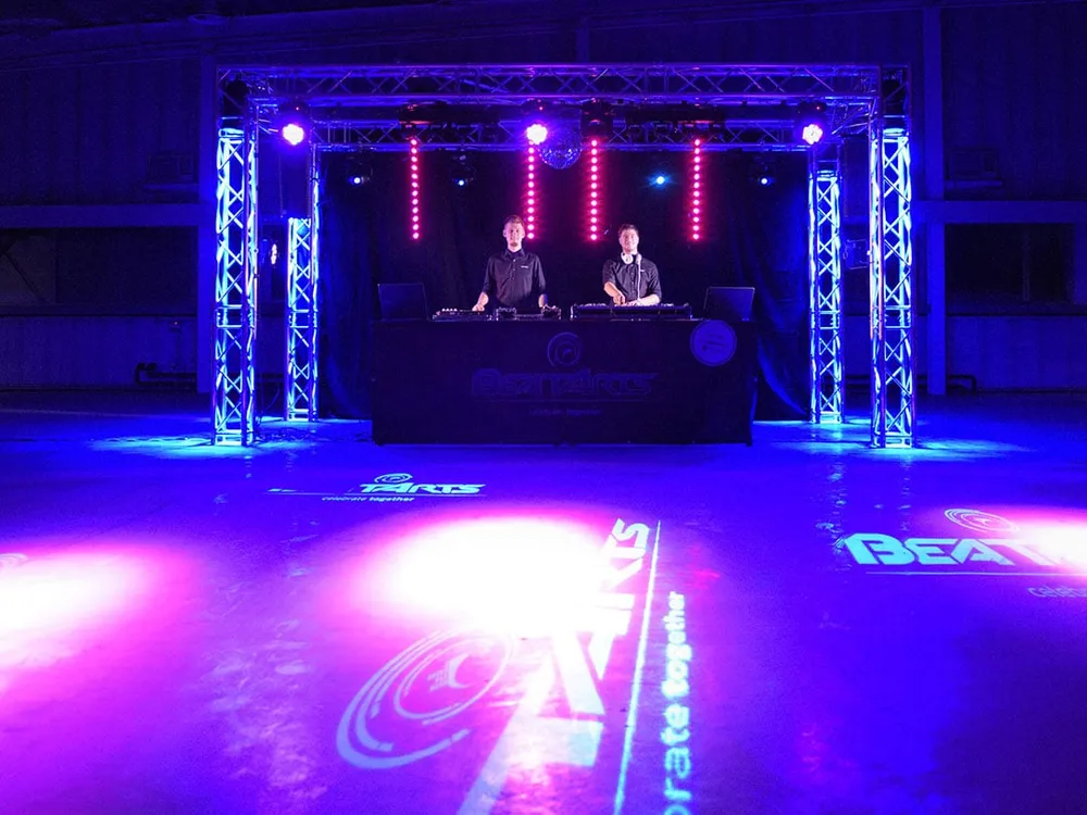 Zwei DJs am BeatArts-DJ-Pult unter einem beleuchteten Trusswürfel, Logo-Projektionen am Boden